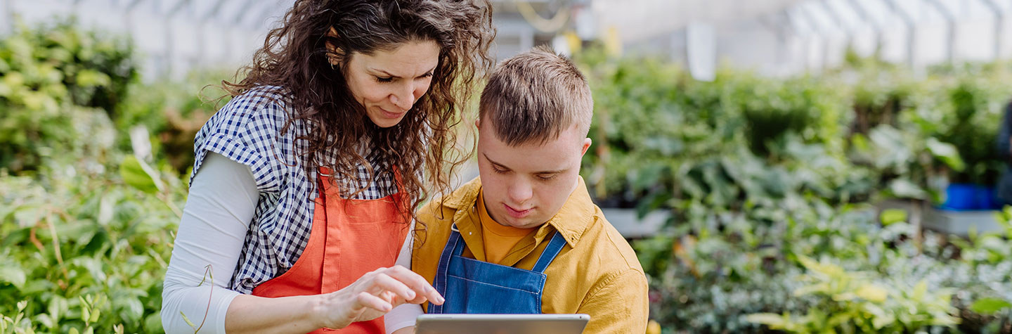 Eine Frau steht neben einem Jungen mit Down-Syndrom, der ein Tablet in der Hand hält, in einem Gewächshaus vor grünen Pflanzen. Beide tragen Arbeitskleidung und schauen konzentriert auf das Tablet.